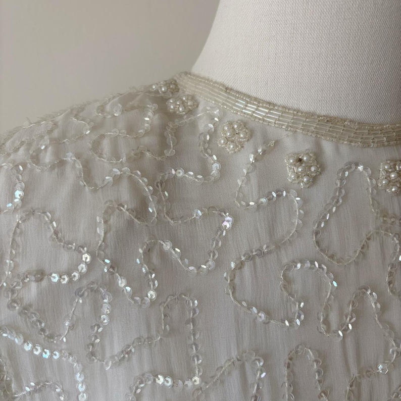 May include: Close-up of a white, vintage top with intricate sequin and bead detailing. The neckline is adorned with delicate beading, and the fabric features flowing, organic patterns of sequins. The overall design is elegant and suitable for formal occasions.