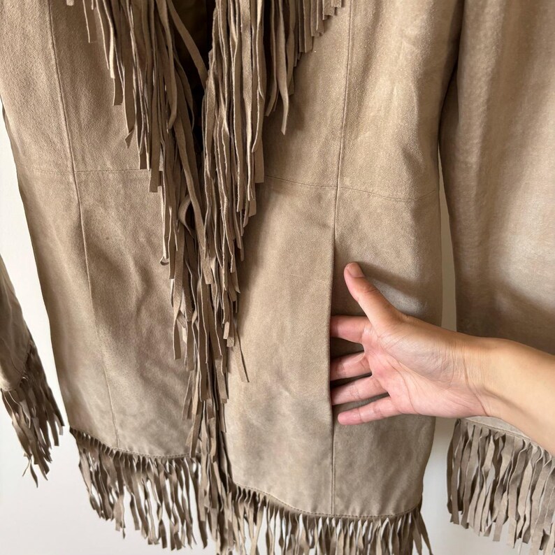 May include: A tan suede jacket with long fringe detailing along the front, hem, and sleeves. The jacket has a classic design with a tailored fit and a pocket. The fringe adds a bohemian flair to the garment.