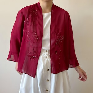 May include: A sheer, open-front burgundy jacket with three-quarter sleeves. The jacket features floral embroidery with sequin accents. The garment is worn over a white top and skirt.