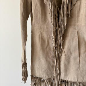 May include: A tan suede jacket with long sleeves and a fringed design. The jacket features fringe along the front, neckline, and sleeves. The jacket is a neutral color and has a classic, bohemian style.