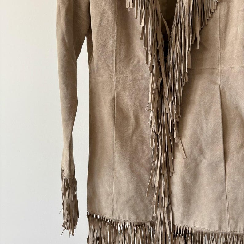 May include: A tan suede jacket with long sleeves and a fringed design. The jacket features fringe along the front, neckline, and sleeves. The jacket is a neutral color and has a classic, bohemian style.