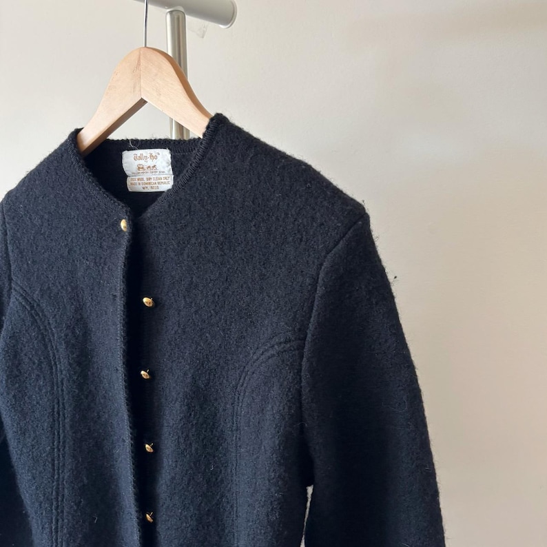 Puede incluir: Chaqueta negra de lana con cuello redondo y botones dorados. La chaqueta est&aacute; colgada de una percha de madera. La chaqueta tiene una superficie texturizada y un corte entallado. La etiqueta es visible en el escote.