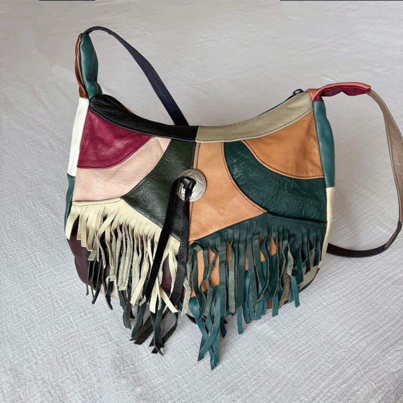 May include: A patchwork leather shoulder bag with a variety of colors including green, tan, black, and burgundy. The bag features a long shoulder strap and decorative fringe along the bottom edge. A silver-toned metal embellishment is on the front.