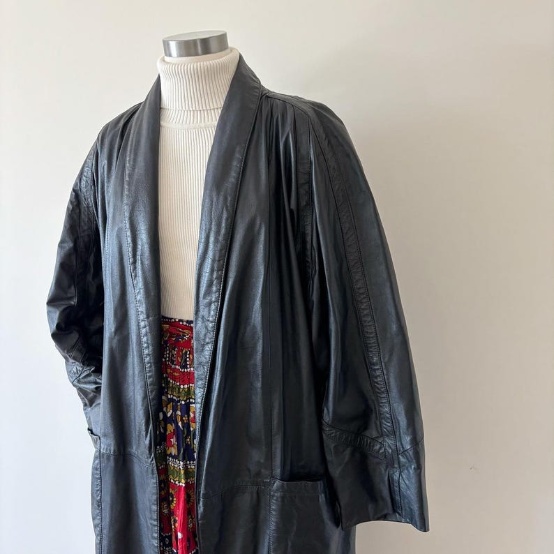 May include: A long, black leather coat with a shawl collar is displayed on a mannequin. The coat has a relaxed fit and pockets. A white turtleneck and a patterned skirt are visible underneath the coat.