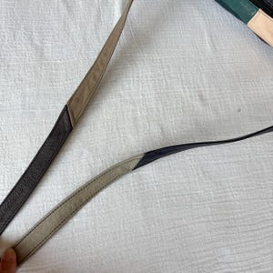 May include: Close-up of two leather bag straps. One strap is dark brown and tan, the other is black and tan. The straps are stitched together with a contrasting thread. The background is a white, textured surface.