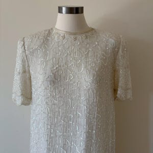 May include: Ivory-colored, short-sleeved dress with intricate sequin and bead detailing. The dress features a round neckline with delicate beadwork and a flowing, textured fabric. The dress is displayed on a mannequin.