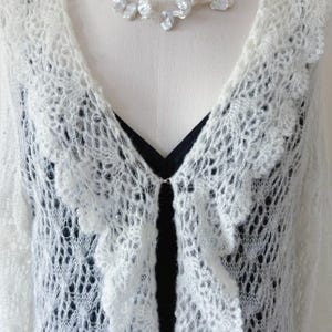 May include: A white, open-front, lacy cardigan with a scalloped edge and a delicate, open weave. The cardigan is paired with a black top and a pearl necklace. The cardigan has a small clasp closure.