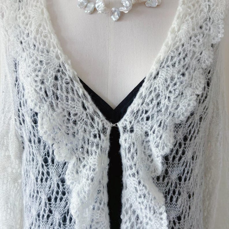 May include: A white, open-front, lacy cardigan with a scalloped edge and a delicate, open weave. The cardigan is paired with a black top and a pearl necklace. The cardigan has a small clasp closure.