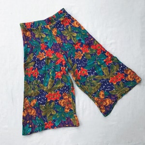 May include: A pair of wide-leg, floral print pants with a blue background. The pants have a high waist and a relaxed fit. The print features a variety of flowers in shades of red, orange, yellow, and green.