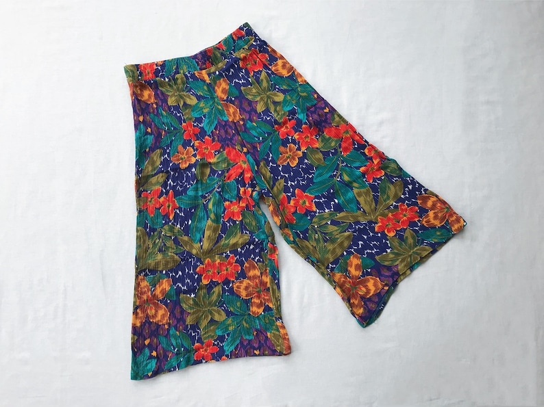 May include: A pair of wide-leg, floral print pants with a blue background. The pants have a high waist and a relaxed fit. The print features a variety of flowers in shades of red, orange, yellow, and green.