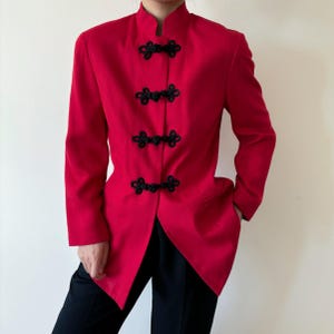 Può includere: Una giacca rosso vivo con colletto alla coreana e chiusure a ranocchia nere. La giacca a maniche lunghe ha una vestibilità sartoriale e un orlo asimmetrico unico. La giacca è abbinata a pantaloni neri, creando un contrasto sorprendente.
