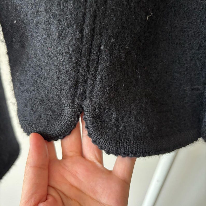 Puede incluir: Primer plano de una chaqueta de lana gris oscuro. La chaqueta tiene un dobladillo festoneado y una superficie texturizada. La tela parece ser un tejido de punto grueso o un material tejido. La chaqueta es probablemente parte de una colecci&oacute;n de oto&ntilde;o o invierno.
