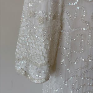 May include: Close-up of a white, embellished garment. The sleeve features a honeycomb pattern of sequins and beads, with additional beaded and sequined details. The fabric appears to be a lightweight material.
