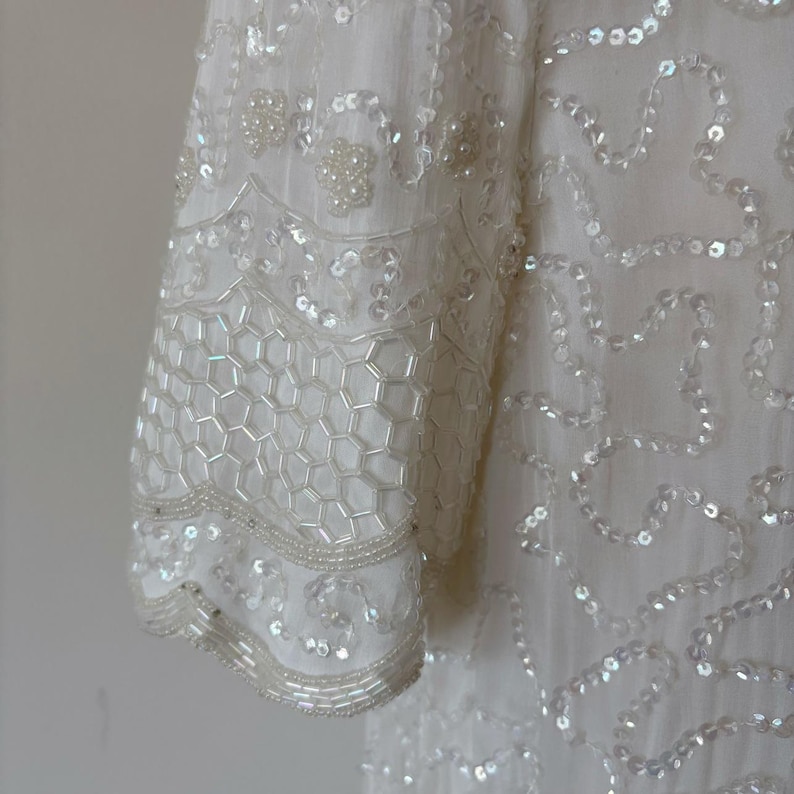 May include: Close-up of a white, embellished garment. The sleeve features a honeycomb pattern of sequins and beads, with additional beaded and sequined details. The fabric appears to be a lightweight material.