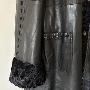 May include: A black leather coat featuring a smooth finish, long sleeves with black fur cuffs, and a large patch pocket on the front. The coat is accented with decorative black stitching along the sleeve seam and the top edge of the pocket.