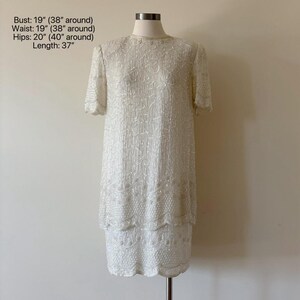 May include: A white, knee-length dress with short sleeves, fully embellished with sequins. The dress is displayed on a mannequin. Measurements: Bust 19 inches, Waist 19 inches, Hips 20 inches, Length 37 inches. The dress has a scalloped hemline.