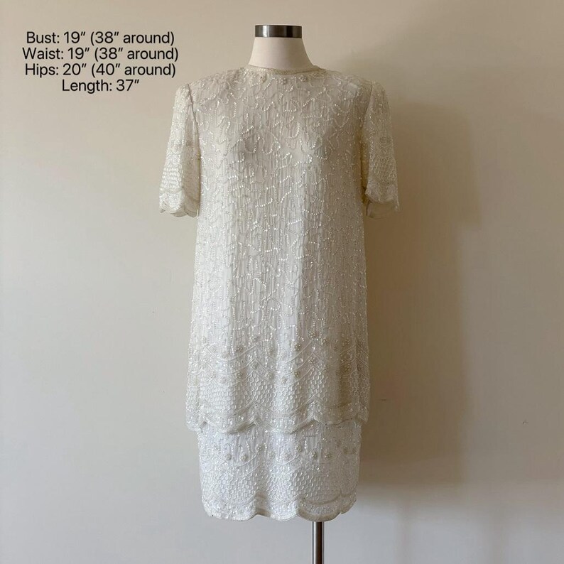 May include: A white, knee-length dress with short sleeves, fully embellished with sequins. The dress is displayed on a mannequin. Measurements: Bust 19 inches, Waist 19 inches, Hips 20 inches, Length 37 inches. The dress has a scalloped hemline.