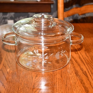 May include: A clear glass saucepan with a matching lid. The pot has two handles and a floral design etched on the side. The pot is sitting on a wooden surface.