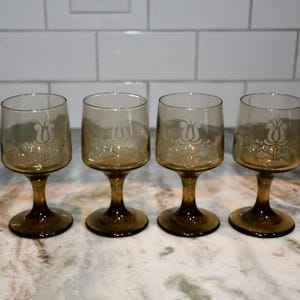 May include: Four amber-colored wine glasses with a tulip design etched on the bowl. The glasses have a rounded bowl and a stem. The glasses are arranged in a row on a marble countertop.