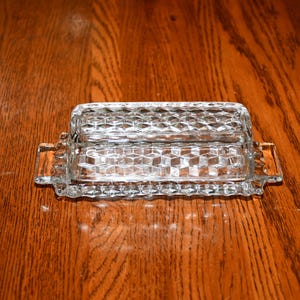 May include: A clear pressed glass butter dish featuring a geometric diamond pattern across the lid and base. The rectangular dish includes two small integrated handles on the sides and a textured rim along the bottom edge of the base.