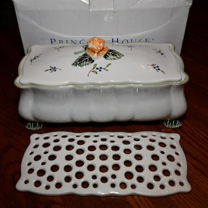 May include: A white ceramic rectangular container with a lid featuring a sculpted orange flower and green leaves. The lid is decorated with small blue berries and green stems. A separate white ceramic grid insert with circular cutouts is included.