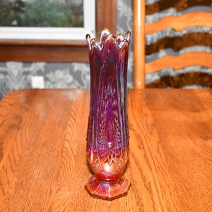 Peut inclure: Un vase en verre violet irisé, de forme élancée, avec un bord évasé et une base hexagonale. Le vase présente un motif texturé et nervuré et mesure environ 25 cm de haut. Le vase est posé sur une table en bois.