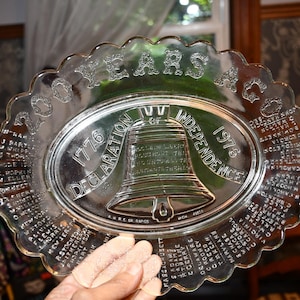 Puede incluir: Plato conmemorativo de vidrio transparente con borde festoneado. El plato presenta un centro ovalado con una imagen de la Campana de la Libertad y el texto "1776 Declaración de Independencia 1976". El borde también tiene texto.