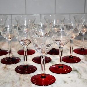 May include: A set of nine clear glass stemmed cocktail glasses with etched floral designs. The glasses have ruby red circular bases. The glasses are arranged on a light-colored countertop.