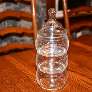 May include: A clear glass stackable canister set consisting of three rounded, tiered sections and a matching lid with a knob handle. Each glass tier features a delicate, etched floral pattern around the exterior surface of the transparent glass.
