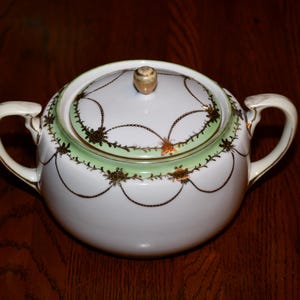 Puede incluir: Azucarero de porcelana blanca con un diseño floral verde y dorado. La tapa tiene un pomo en la parte superior.