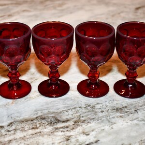 May include: Four ruby red wine glasses with a decorative pattern. The glasses have a round base, a stem, and a bowl. The glasses are arranged in a row on a marble countertop. The glasses are empty.