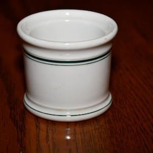 May include: White ceramic napkin ring with a cylindrical shape. The ring features two thin, dark green stripes around the body. The top and bottom edges are slightly flared. The napkin ring is set on a dark brown wooden surface.