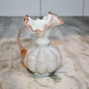 May include: A white glass pitcher with a ruffled rim and a handle. The pitcher has a bulbous body with a series of vertical ridges. The handle and rim have a pinkish-orange tint.
