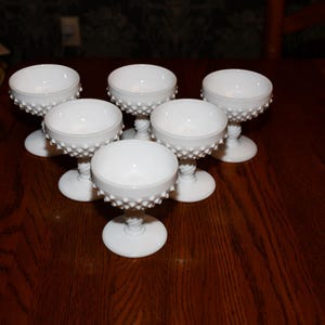 May include: Six white hobnail glass dessert or champagne glasses. Each glass features a round bowl atop a stem with a textured, hobnail design. The glasses are arranged on a dark wooden surface.