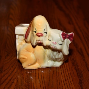 May include: A ceramic figurine featuring two dogs. One dog is tan with red accents, and the other is white with a red bow. The dogs are nestled together, with the white dog resting its head on the other. The figurine is a decorative item.