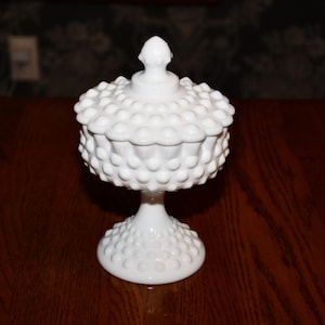 Könnte beinhalten: Eine weiße Hobnail-Bonbonniere mit Deckel. Die Schale hat einen Sockel und einen runden Körper, der mit kleinen, erhabenen Noppen bedeckt ist. Der Deckel ist ebenfalls mit Noppen bedeckt und hat eine kleine, spitze Spitze. Die Schale steht auf einer Holzoberfläche.
