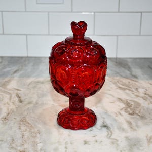 May include: A ruby red glass candy dish with a lid. The dish has a pedestal base and is decorated with a star and moon pattern. The lid is topped with a heart-shaped finial. A decorative piece for the home.