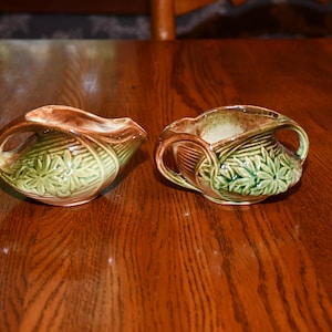 Könnte beinhalten: Zwei Keramik-Sahnekännchen mit Griffen. Die Kännchen haben einen braunen Rand und Griff, mit einem grünen Blumendesign auf dem Körper. Die Kännchen stehen auf einer Holzoberfläche.