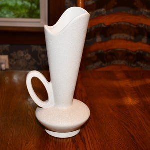 May include: A white ceramic vase with a textured, matte finish. The vase features a wide, rounded base that tapers into a tall, slender neck with an asymmetrical, curved rim. A single, circular handle is attached to the side of the neck.