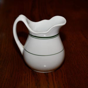 May include: A white ceramic pitcher with a curved handle and a scalloped rim. The pitcher has two dark green bands around the body. The pitcher is sitting on a dark brown wooden surface.
