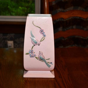 Puede incluir: Un jarrón de cerámica rosa con base rectangular y parte superior acampanada. El jarrón está decorado con dos pájaros azules y turquesas posados en una rama con flores azules. El jarrón está sobre una mesa de madera.