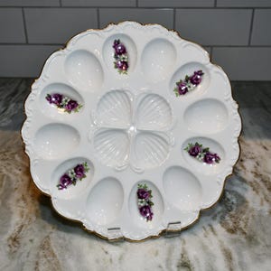 May include: A white ceramic egg serving platter with a scalloped edge and gold trim. The platter features twelve oval indentations for eggs, with four central shell-shaped wells. Five of the egg indentations are decorated with purple rose floral decals.