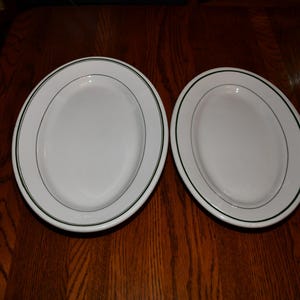 May include: Two white oval serving platters with a green trim on a dark wooden surface. The platters are empty and appear to be made of ceramic or porcelain. The plates are side by side.