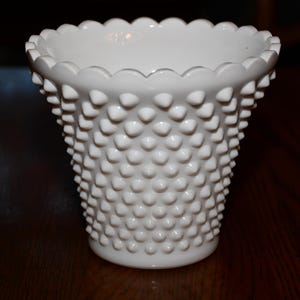 Puede incluir: Un jarrón blanco hobnail con un borde festoneado. El jarrón tiene una superficie texturizada de pequeñas protuberancias redondeadas. El jarrón está hecho de cerámica o vidrio y está sobre una superficie de madera oscura. El jarrón está vacío.