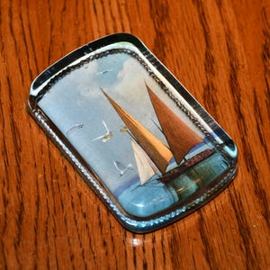 Puede incluir: Un pisapapeles rectangular de vidrio con una escena náutica. La imagen representa veleros con velas marrones sobre un mar azul bajo un cielo azul claro. El pisapapeles tiene un borde plateado y está sobre una superficie de madera.