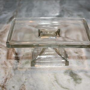 Könnte beinhalten: Ein klarer, quadratischer Kuchenständer aus Glas. Er hat eine rechteckige Oberseite und einen Sockel. Der Ständer besteht aus dickem Glas und hat ein schlichtes Design. Geeignet für die Präsentation von Kuchen oder anderen Desserts. Misst ca. 20 cm im Quadrat.