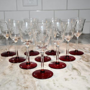 May include: Nine clear wine glasses with etched floral designs. Each glass has a long stem and a round, deep red base. The glasses are arranged on a marble countertop.