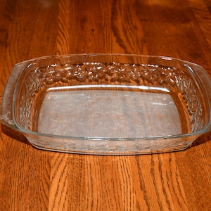 May include: A clear, rectangular glass baking dish with a decorative floral pattern around the rim. The dish has a rectangular shape with slightly curved sides and handles. It sits on a wooden surface.
