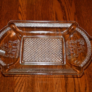 May include: Clear glass serving tray with a rectangular center and decorative edges. The tray features embossed designs on the sides and a textured pattern in the center. The tray is made of clear glass.