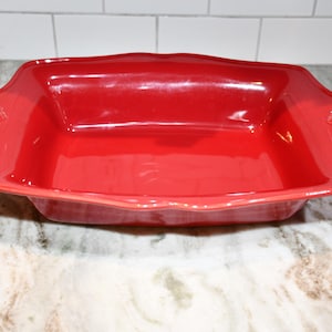 May include: A rectangular, red ceramic baking dish with decorative handles. The dish has a glossy finish and is on a marble countertop. The baking dish is designed for cooking and serving food.